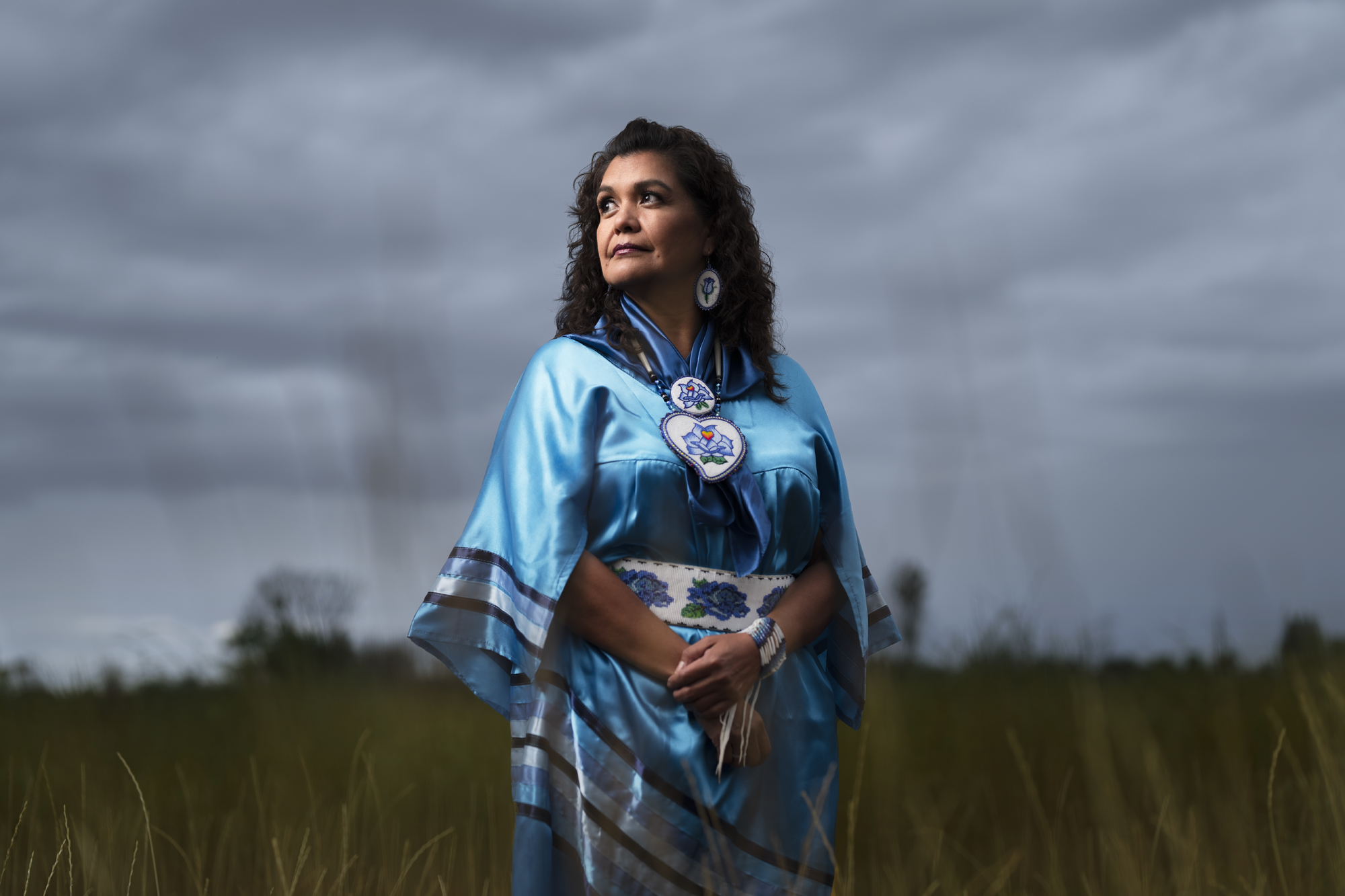 woman in regalia looking off into the distance with tall grass behind her